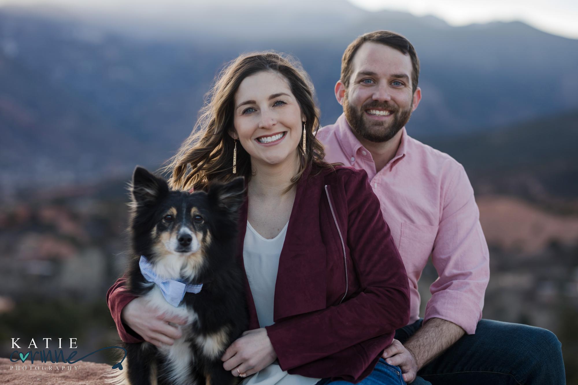 engaged couple holds their dog and looks at camera