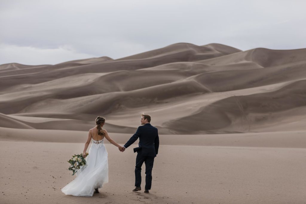 bride and groom hold hands on sand dunes