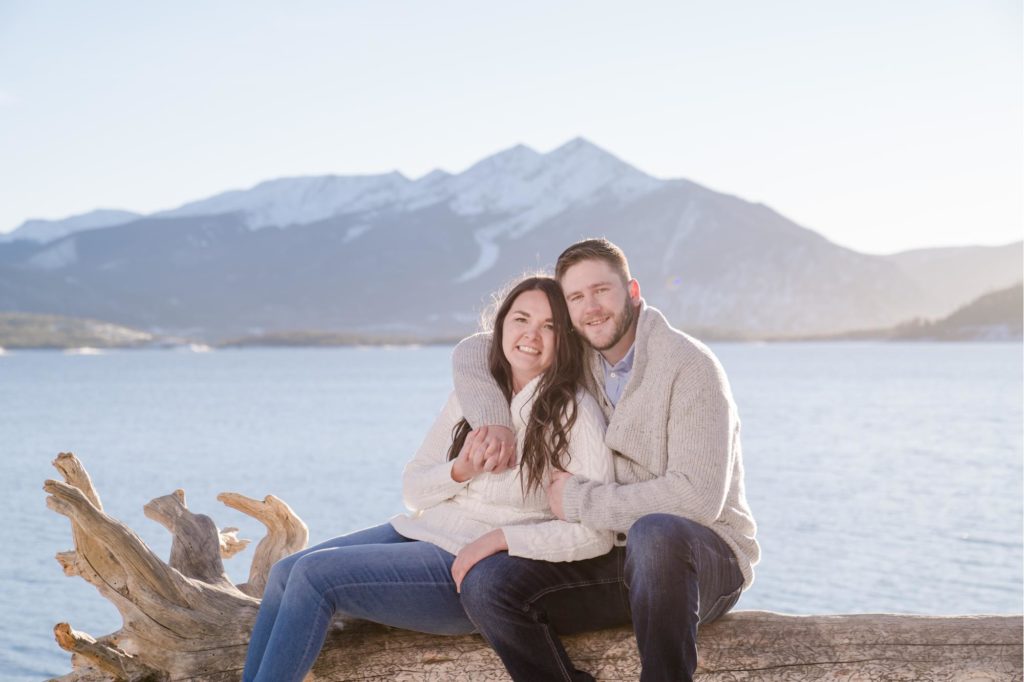 engaged couple poses on log at lake